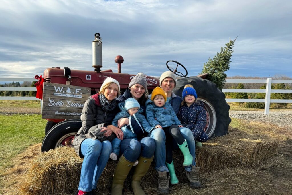 A family sitting on hay bales in front of an old red tractor at a Christmas tree farm selfie station.