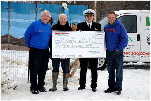 Four individuals, including a man in a US Navy uniform, stand in the snow holding a large novelty check for "$21,000".