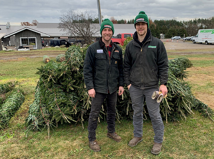 Two men in green hats standing next to a large pile of donated Christmas trees.