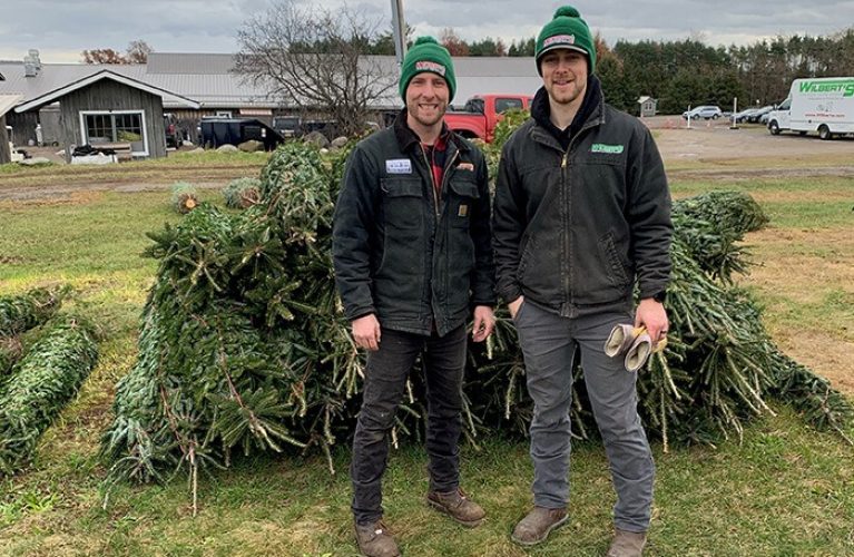 Two men in green hats standing next to a large pile of donated Christmas trees.