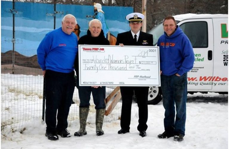 Four individuals, including a man in a US Navy uniform, stand in the snow holding a large novelty check for "$21,000".
